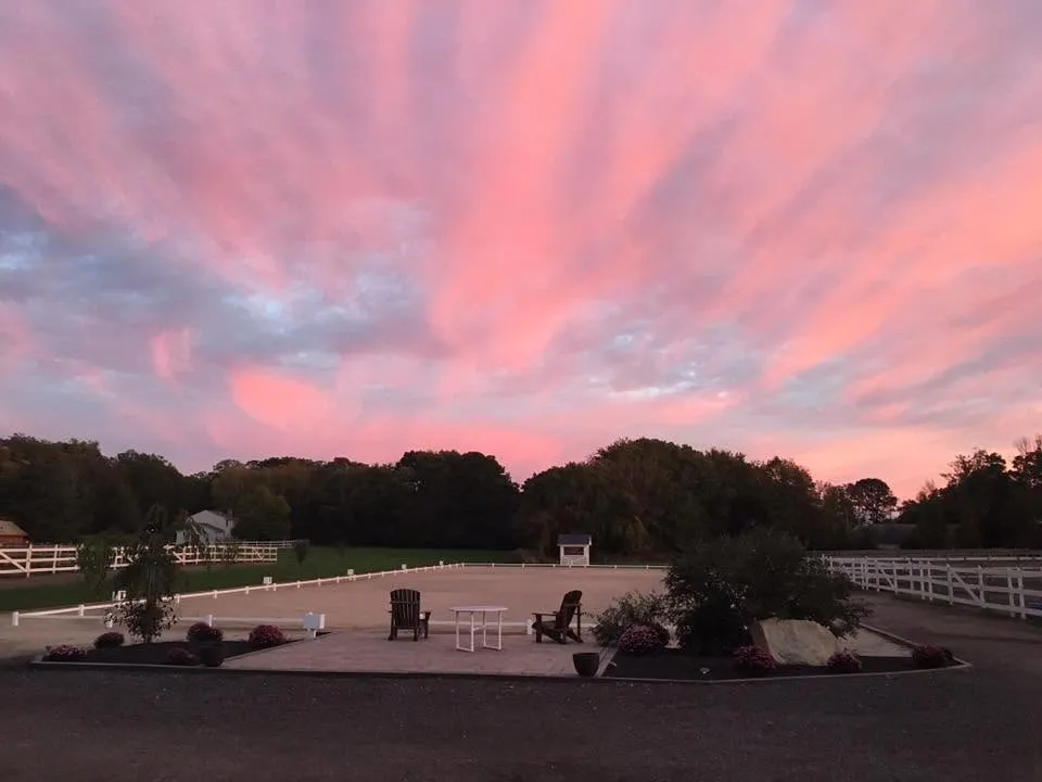 A pink sunrise over the outdoor dressage arena at Lepore Farm, with Adirondack chairs on the patio